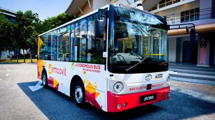 A compact white, red, and yellow autonomous electric bus with "eMoovit" and "Bas Berautonomi" decals parked in front of a modern civic building.