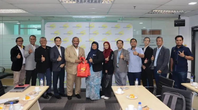 A group of corporate and government representatives standing together giving thumbs-up gestures inside a meeting room, with two leaders holding a gift bag in the centre.
