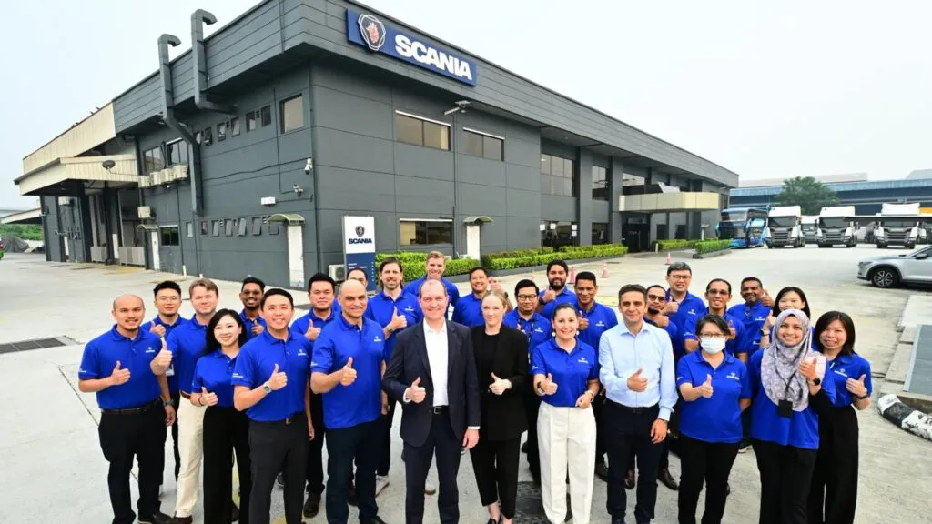 Dr Michael Jackstein, Carolin Riethus, and Scania Southeast Asia staff wearing blue shirts giving thumbs-up gestures outdoors in front of the Scania Bukit Jelutong headquarters building.
