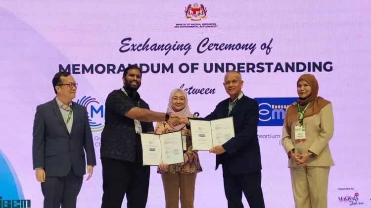 Representatives from MGTC and Konsortium E-Mutiara holding open document folders during a formal MoU exchange ceremony on stage in front of a Ministry backdrop.