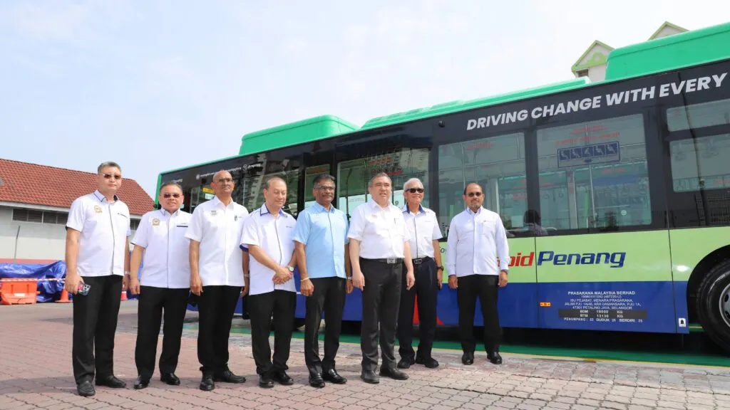A group of corporate and ministry officials standing in a row in front of a green and blue Rapid Penang electric bus bearing the slogan Driving Change With Every Journey.