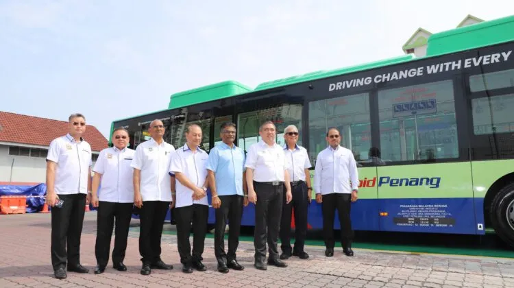 A group of corporate and ministry officials standing in a row in front of a green and blue Rapid Penang electric bus bearing the slogan Driving Change With Every Journey.
