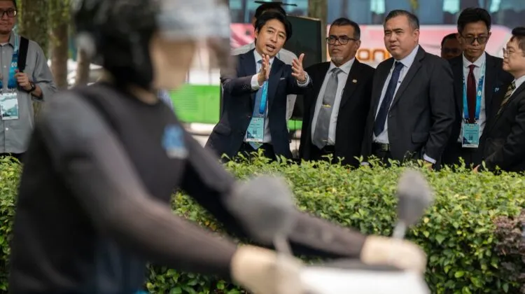 Transport Minister Anthony Loke in a suit watching an outdoor live demonstration of a test dummy riding a motorcycle behind green hedges.